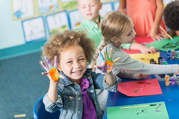 A cute little girl in art class in preschool or kindergarten.  She is sitting at a table with other children painting pictures.  She is looking at the camera, smiling, holding up her hands covered in paint.