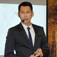Patric Chan speaking at a presentation dressed in a black suit and tie with a white shirt.