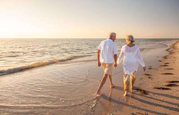 Happy senior man and woman old retired couple walking and holding hands on a beach at sunset