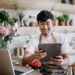 Asian male florist, owner of small business flower shop, using digital tablet while working on laptop against flowers and plants. Checking stocks, taking customer orders, selling products online. Daily routine of running a small business with technology