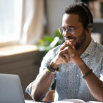 Happy millennial african american man in glasses wearing headphones, enjoying watching educational webinar on laptop. Smiling young mixed race businessman holding video call with clients partners.